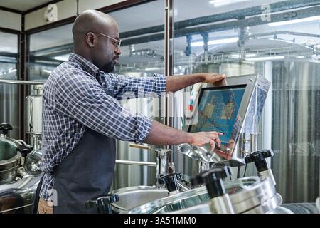 African American engineer operating equipment at a brewery, monitored on a computer screen. An industrial setting highlighting engineering, manufacturing, and beer production. Ideal for tech in food and beverage, factory work, and skilled trades. Stock Photo