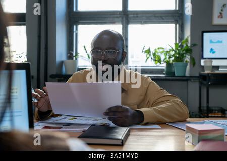 Serious Black businessman holding financial papers sits opposite an Asian businesswoman in a bright office. The pair appear engaged in a strategic discussion about finance or startup planning. This image highlights diversity in the workplace and executive collaboration. Stock Photo