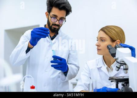 Indian male and caucasian female scientists using pipette, test tube, and microscope researching medical sample together in laboratory Stock Photo