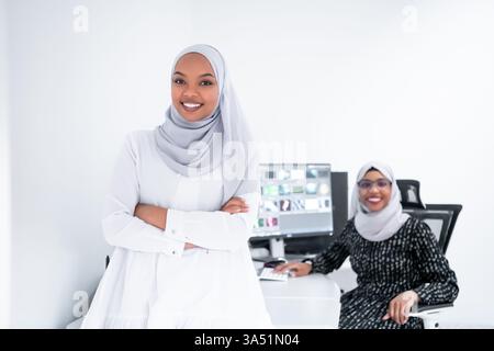 Two young Afro-American Muslim businesswomen in a bright, modern office with a large screen. The scene conveys collaboration, creativity, and teamwork in a diverse workplace. Suitable for startup culture, meetings, and women in leadership visuals. Stock Photo
