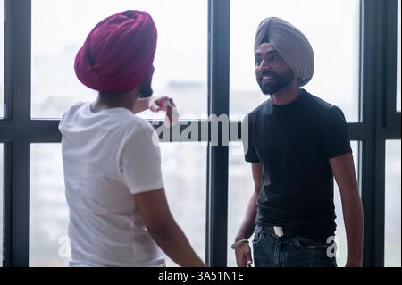 Two Indian men wearing turbans talking near a window in a home setting. The scene reflects everyday conversation and cultural diversity, suitable for lifestyle or community features. Stock Photo