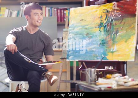 Chinese artist painting in his studio, surrounded by easel, brushes and colorful canvases. The portrait highlights creativity and the art process in a modern studio setting. Suitable for features on artists, studios and Chinese art culture. Stock Photo