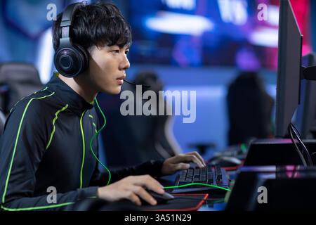 Focused Chinese man wearing a black jacket and headphones works at a computer in an internet cafe. A strong choice for esports, gaming and technology themed projects featuring a young adult in an Asian setting. Stock Photo