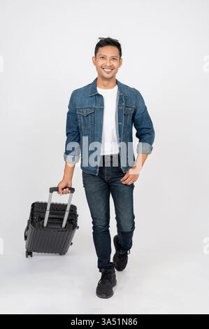 Smiling Asian man in a denim jacket with luggage stands confidently against a white studio backdrop. This portrait suits travel and luggage themes, from arrivals to holiday planning. Stock Photo