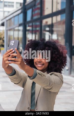 Happy African American woman in a business suit takes a cheerful selfie on a city street before work. The urban scene highlights professionalism, confidence, and a modern workplace lifestyle. Stock Photo
