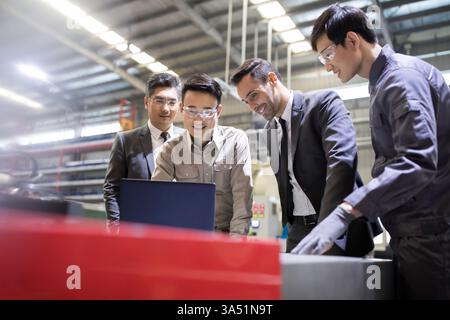 Smiling Chinese businessmen and mechanical engineers stand in a factory, looking at a laptop screen. The image showcases collaboration and technical expertise in manufacturing settings. Ideal for industrial and engineering collaboration themes. Stock Photo