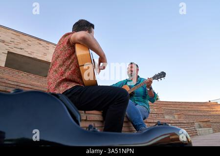 Two male musicians play acoustic guitars on city steps, captured from below in an urban street scene. The cheerful duo performs live outdoors, conveying friendship and talent. Ideal for street performance, urban lifestyle, and music themes. Stock Photo