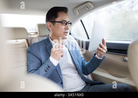 Chinese businessman in a private car uses a digital tablet for work on the go. This in-car technology scene is ideal for content about mobile business and connectivity. Stock Photo