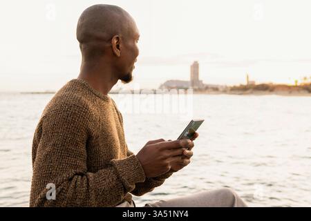 Afro-Latino man looking away while holding a smartphone on a sunny beach. This relaxed seaside portrait conveys connectivity and leisure, ideal for travel, lifestyle, and mobile technology themes. Great for beach lifestyle campaigns with natural daylight and calm sea backdrop. Stock Photo
