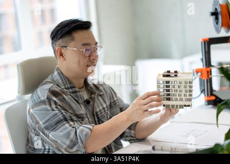 An Asian man examines a 3D printed house model in a bright office within a creative lab. The scene highlights 3D printing technology, architectural modeling, and collaborative work among designers and engineers. Perfect for features on innovation, design studios, and modern manufacturing. Stock Photo