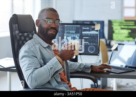 Portrait of an African American software engineer at a modern desk, coffee cup in hand and looking at the camera with copy space. This tech and business image works well for information technology, software development, and professional branding. Great for corporate communications and tech blogs. Stock Photo