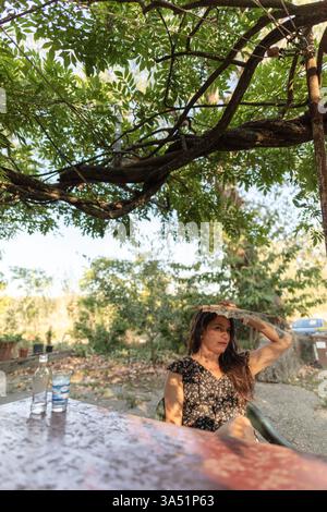 Hispanic woman holds a fan above her head to shade from the sun, seated at a table under a tree in a rural outdoor setting. This relaxed lifestyle scene suits outdoor living, travel, and rural lifestyle campaigns. Stock Photo
