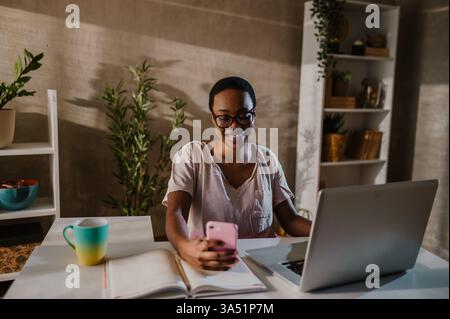 Smiling young African American woman uses a laptop and a smartphone, engaged in online chats or shopping. A modern home office scene with copy space for text or graphics. Ideal for telecommuting, remote work, and digital lifestyle themes. Stock Photo