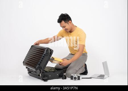 Smiling Asian man opens his luggage and sits on the floor against a white studio backdrop. This travel-preparation image works well for lifestyle and packing themes. Stock Photo