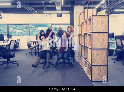 Multiracial startup team of software developers having fun racing office chairs in the office. The diverse group show energy, teamwork and a playful corporate culture. Stock Photo