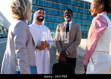 A diverse group of business professionals meets outdoors in an urban setting for a group discussion and team bonding. The scene highlights multicultural collaboration and networking against a modern city backdrop. Stock Photo