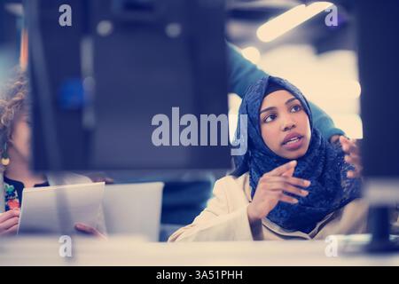 Professional Black Muslim woman wearing a hijab sits at a conference table in a modern office, raising her hands as she contributes to a meeting near a laptop. This scene highlights diversity in the workplace and collaborative teamwork with technology. Ideal for corporate communications, inclusive workplaces, and business presentations. Stock Photo