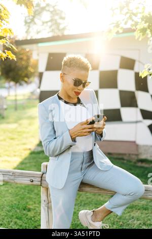 Hispanic woman using smartphone while sitting on a railing in a sunny park. This bright outdoor scene emphasizes mobile connectivity, casual fashion, and park life, perfect for lifestyle and social media campaigns. Suitable for fashion, mobile apps, and outdoor advertising. Stock Photo