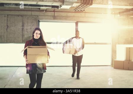 Happy multiethnic business team moving into a new startup office, carrying cardboard boxes with sunlight streaming through windows. The scene emphasizes teamwork, relocation, and a dynamic startup atmosphere. Great for articles about office moves, startups, or collaborative work cultures. Stock Photo