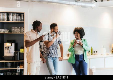 Three colleagues share a coffee break in a modern coworking kitchen, enjoying a casual chat. The diverse team atmosphere highlights collaboration, informal work culture, and work-life balance in an urban office setting. Stock Photo
