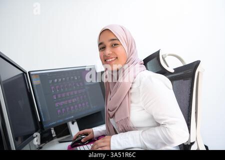 Female Arabic creative professional working at a home office with a dual-monitor setup, wearing a hijab. A modern work-from-home scene that highlights entrepreneurship, technology, and focused productivity in a contemporary interior. Stock Photo