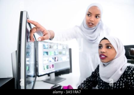 Two young Afro-American Muslim businesswomen in a bright modern office, wearing scarves, collaborate near a large screen. The scene conveys teamwork, creativity, and inclusive leadership in a contemporary workplace. Great for features on diversity in business and female entrepreneurship. Stock Photo