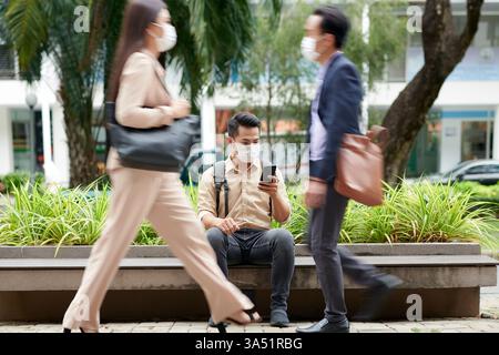 Asian man wearing face mask using smartphone sitting on bench in city streets during daytime Stock Photo