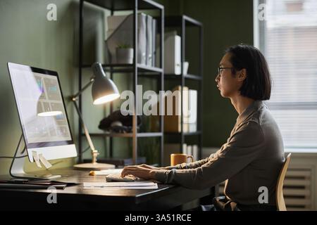 Side view portrait of an Asian software designer working late at a home office. The scene emphasizes concentration, technology, and creativity in a modern workplace. Copy space is available for headlines and messaging. Stock Photo
