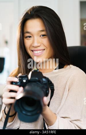 Smiling Asian woman sits in a modern office, holding a camera as part of her work. This portrait conveys professionalism and creativity in a contemporary workplace. Stock Photo
