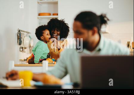 A happy African American family in the kitchen, with the mother feeding her son while the father finishes remote work on a laptop. A warm, everyday family moment suitable for home life, parenting, and work-life balance themes. Stock Photo