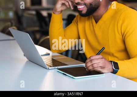 Black man working on a laptop with a tablet in an office setting. Great for tech, remote work, digital collaboration, and software development stories. Stock Photo