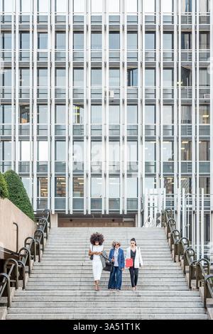 Three businesswomen, Hispanic and Caucasian, walking downstairs in a city building during daytime. The scene conveys professional teamwork in a modern urban workplace. Stock Photo