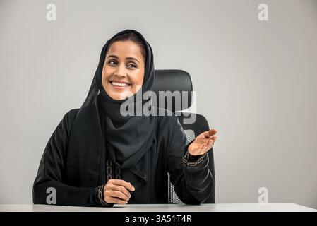 Smiling Indian woman sits at an office desk, raising an open hand in a friendly gesture. A welcoming corporate portrait suitable for business communications and workplace positivity. Useful for lifestyle and professional branding imagery. Stock Photo