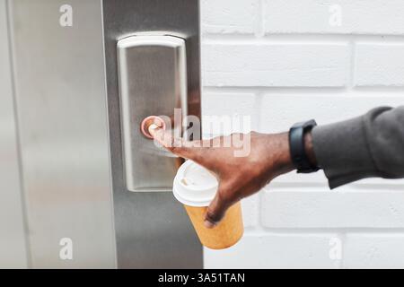 Close-up of a young African American man pushing an elevator button in an office setting, coffee cup in hand. The shot includes copy space for branding or messaging, perfect for business and workplace themes. Great for corporate communications, productivity and modern office lifestyle. Stock Photo
