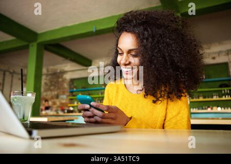 Smiling Black woman with curly hair using a smartphone at a cafe table with a laptop. Casual, modern urban lifestyle scene perfect for topics on mobile tech, remote work, and cafe culture. Great for brands targeting diverse young professionals and digital workflows. Stock Photo