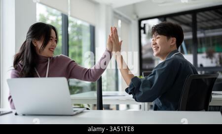 Smiling Asian male and female office workers giving high five to each other sitting at desks in office Stock Photo