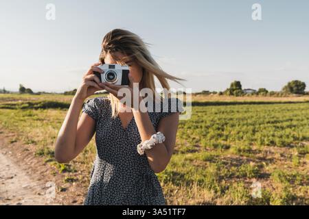 Hispanic woman stands on a sunny countryside road, using a retro camera to capture travel moments. The image evokes nostalgia, outdoor photography, and wanderlust. Great for travel, photography, and lifestyle storytelling. Stock Photo