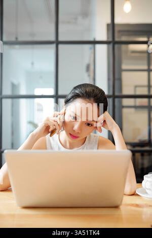 Focused female entrepreneur making a phone call while reviewing reports on a laptop. This professional scene supports business, women in leadership, and digital collaboration themes. Stock Photo