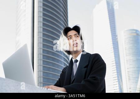 Asian man in smart casual works on a netbook with city buildings in the background. A modern urban scene suitable for tech, startups, and business communications. Great for corporate blogs, online work culture, and remote-working themes. Stock Photo