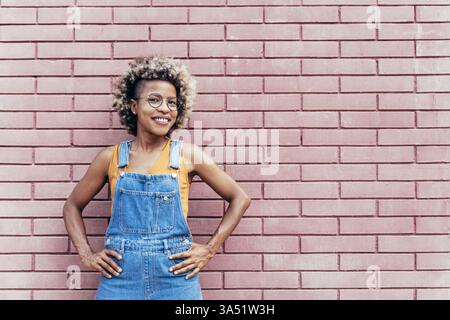 Smiling Black woman standing with hands on hips against a brick wall outdoors. She exudes confidence and casual style in an urban setting. A versatile image for empowerment, fashion, and everyday moment campaigns. Stock Photo