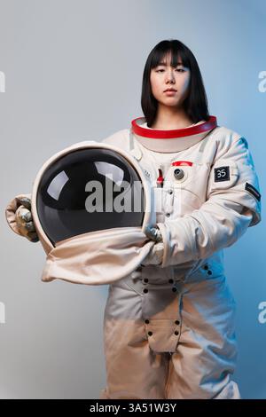 Serious Asian woman wearing spacesuit holding helmet standing with blue light reflection against white wall Stock Photo