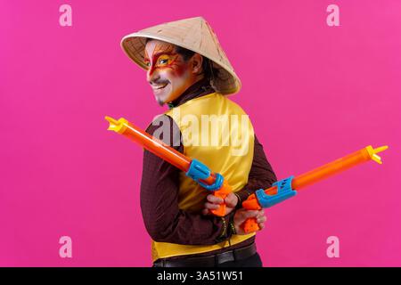 Colorful studio portrait of a clown with white facial makeup against a pink backdrop. The image features playful props including toy guns and a Chinese hat, contributing to a carnival and humor mood. Ideal for entertainment, party, circus and comedy themes in ads or editorial content. Stock Photo