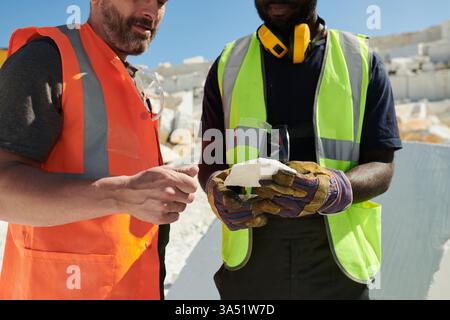 Midsection shot of two intercultural workers in uniform discussing a block of marble at a quarry. The scene highlights teamwork and professionalism in an industrial setting. A strong choice for articles about diverse teams, construction, or the stone industry. Stock Photo