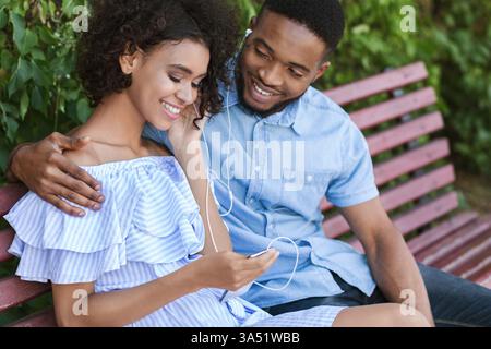 Young couple sits on a park bench while using a smartphone, sharing a light moment outdoors. Perfect for lifestyle, dating, and social campaigns about connection and modern tech. Stock Photo