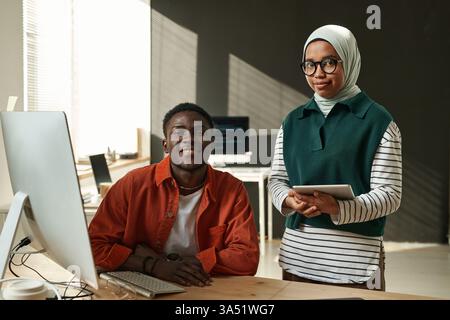 Black male software developer sitting at table with laptop beside Mixed race Muslim female coworker holding tablet standing in office Stock Photo