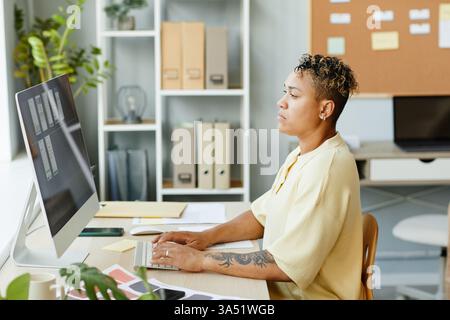 Side view of a tattooed Black woman using a computer in an office while designing a mobile app interface. Focused and creative, she works on UI and UX development. Great for technology, software design, and women in tech themes. Stock Photo