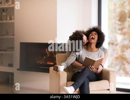 Young multiethnic couple hugging in front of a fireplace at home while reading a book. Cozy autumn interior scene with warm lighting and a relaxed, intimate mood. Ideal for home lifestyle, family moments, and autumn or holiday storytelling. Stock Photo