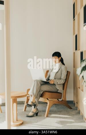 A focused professional Japanese woman dressed in a light gray suit engages with her laptop while seated comfortably. The soft lighting creates a seren Stock Photo