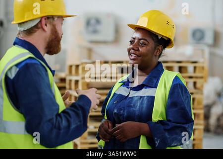 Two young intercultural engineers in workwear discuss features of new machinery in a meeting. They stand in front of the equipment, highlighting teamwork and technical expertise in an industrial setting. Ideal for engineering, manufacturing and diversity-themed campaigns. Stock Photo