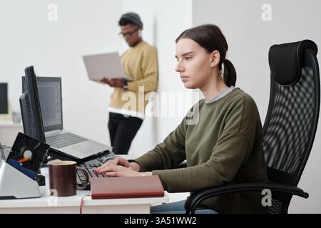 Mixed race woman works on a computer at a table while a Black male colleague leans on the wall nearby with a laptop. The image represents diverse, intercultural teamwork in a contemporary office. Ideal for articles on collaboration, leadership, and tech teams. Stock Photo